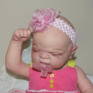 May include: A pink and white headband with a flower accent is worn by a baby doll with closed eyes. The doll is wearing a pink and yellow dress and has a pacifier in its mouth.