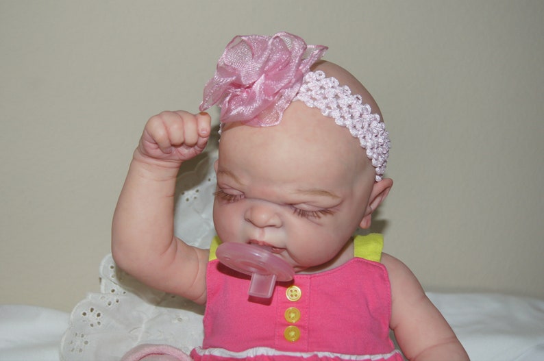 May include: A pink and white headband with a flower accent is worn by a baby doll with closed eyes. The doll is wearing a pink and yellow dress and has a pacifier in its mouth.