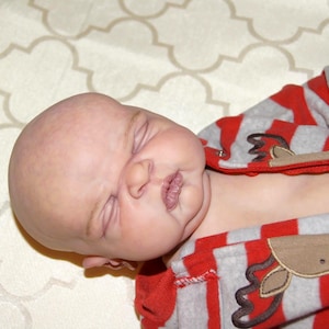 May include: A sleeping baby wearing a red and gray striped onesie with a reindeer applique. The onesie has long sleeves and a zipper. The baby is lying on a patterned surface.