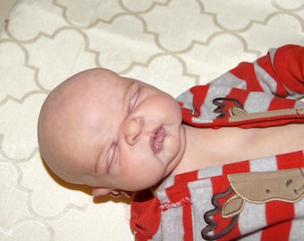 May include: A sleeping baby wearing a red and gray striped onesie with a reindeer applique. The onesie has long sleeves and a zipper. The baby is lying on a patterned surface.
