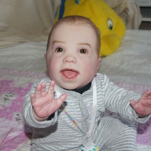 Puede incluir: Un mu&ntilde;eco de beb&eacute; con ojos azules y una amplia sonrisa lleva un atuendo a rayas grises y blancas con una cinta blanca alrededor del cuello. El mu&ntilde;eco est&aacute; sentado en una manta estampada rosa y blanca.