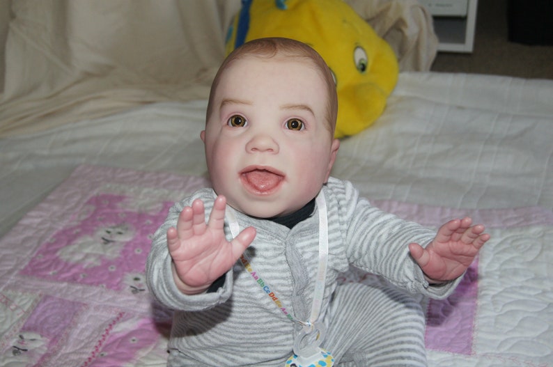 Puede incluir: Un mu&ntilde;eco de beb&eacute; con ojos azules y una amplia sonrisa lleva un atuendo a rayas grises y blancas con una cinta blanca alrededor del cuello. El mu&ntilde;eco est&aacute; sentado en una manta estampada rosa y blanca.