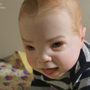 Puede incluir: Primer plano de una mu&ntilde;eca beb&eacute; con cabello casta&ntilde;o claro y ojos marrones. La mu&ntilde;eca lleva una camisa a rayas blancas y negras y mira a la c&aacute;mara con una expresi&oacute;n juguetona.