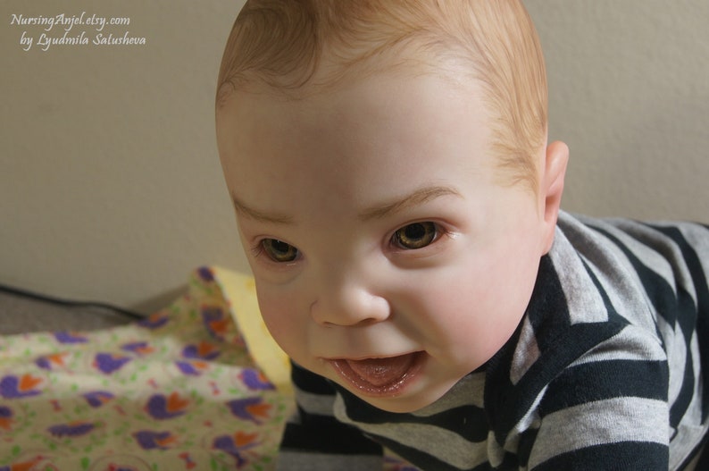 Puede incluir: Primer plano de una mu&ntilde;eca beb&eacute; con cabello casta&ntilde;o claro y ojos marrones. La mu&ntilde;eca lleva una camisa a rayas blancas y negras y mira a la c&aacute;mara con una expresi&oacute;n juguetona.