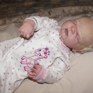 May include: A realistic baby doll wearing a white onesie with pink heart patterns and a giraffe design. The doll has blonde, curly hair and is lying on a light-colored surface.