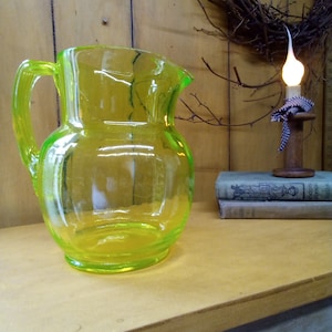 Op de afbeelding: Een heldergele glazen kan met een rond lichaam en een handvat. De kan staat op een houten oppervlak.
