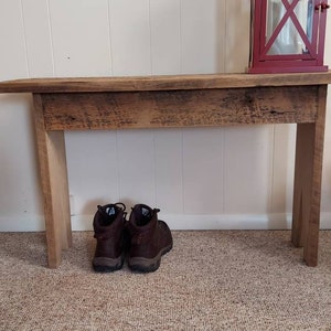 May include: A rustic wooden bench with a distressed finish, featuring a burgundy lantern on top and a pair of brown hiking boots beneath. The bench is made of light-colored wood and stands against a white wall.