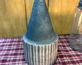 Puede incluir: Un objeto decorativo con forma de cono sobre una base cilíndrica. El cono es de color azul oscuro texturizado, mientras que la base presenta rayas verticales blancas y azules. La superficie del objeto parece desgastada con un acabado mate.