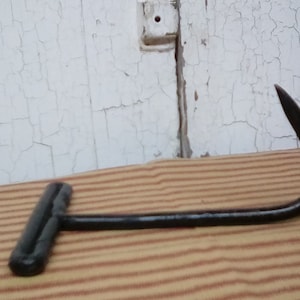 Puede incluir: Un gancho de metal negro con un mango en forma de T. El gancho es curvo y tiene una punta afilada. Está sobre una superficie rayada.