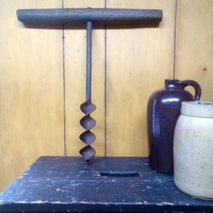 Könnte beinhalten: Ein antiker Schneckenbohrer mit Holzgriff und Spiralbohrer, der auf einer dunklen Holzkiste ruht. Eine dunkelbraune Kanne und ein cremefarbenes Glas mit Deckel befinden sich im Hintergrund, vor einer gelben Holzwand.