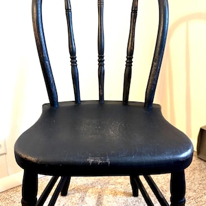 May include: A black wooden chair with a curved backrest, three vertical spindles, and a contoured seat. The finish shows signs of wear with visible wood grain and light distressing along the edges of the seat and the back frame structure.