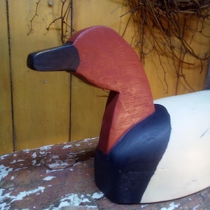 May include: A wooden duck figurine with a red head and neck, a black beak and collar, and a white body. The duck is painted with a distressed finish, giving it a rustic appearance. The figurine is placed on a weathered wooden surface.