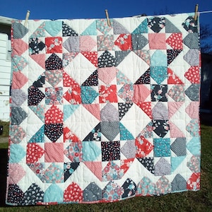 May include: A patchwork quilt with a geometric pattern in shades of pink, blue, gray, and white. The quilt is made of various fabrics with floral and geometric prints.