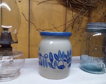 May include: A cylindrical stoneware crock with a light beige base and a thick blue rim. The exterior features a hand-painted blue floral and vine pattern encircling the middle section. The vessel has a smooth, glazed finish and a wide opening.