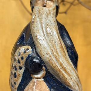 May include: A ceramic figurine of an elderly man with a long, flowing beard and a hat. The figure is dressed in a dark blue robe with a lantern in his hand. The beard and hat have a speckled design.