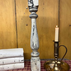 May include: A tall, weathered wooden baluster with chipped white paint revealing dark wood underneath, positioned next to a stack of three white books and a brass candle holder with a lit white taper candle on a red and tan plaid surface.
