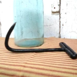 May include: A light blue glass jar with a threaded metal lid positioned behind a black metal T-shaped hay hook. The jar is cylindrical with a smooth surface, while the hook features a curved, pointed end and a horizontal handle grip.