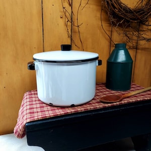 Może przedstawiać: Biały emaliowany garnek z czarną obwódką i uchwytami, stojący na czerwono-brązowym obrusie w kratkę. Garnek ma czarną pokrywkę i stoi na czarnym drewnianym stołku.