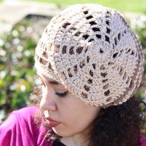 May include: A woman wearing a crocheted beige hat with a lacy design. She is wearing a pink scarf and a black shirt with a patterned design.