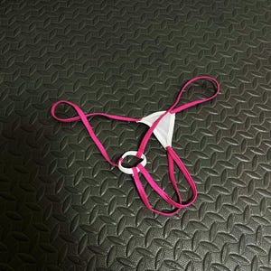 Pode incluir: Uma tanga branca e rosa. A roupa interior apresenta um painel frontal triangular branco, alças rosa e um detalhe de anel branco. O design é minimalista.