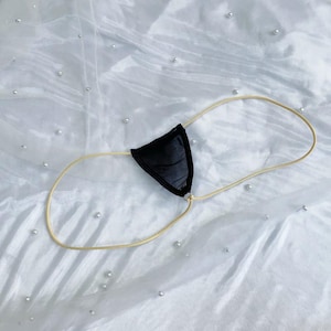 Puede incluir: Lencería negra con un diseño triangular y finas correas beige. La prenda está hecha de una tela oscura y lisa y está diseñada para usarse alrededor de la cintura. El fondo es una superficie blanca texturizada.