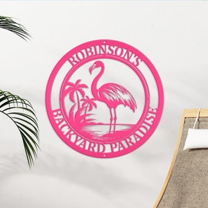 Puede incluir: Señal de metal rosa con el texto "Robinson's Backyard Paradise" y una silueta de flamenco con palmeras en el fondo.