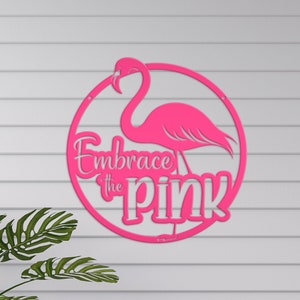 Könnte beinhalten: Rosa-farbene Metall-Flamingo-Wandkunst mit dem Text "Embrace the Pink" in einem Kreis. Die Kunst ist an einer weißen Wand mit einer grünen Pflanze und einer gestreiften Couch im Vordergrund montiert.
