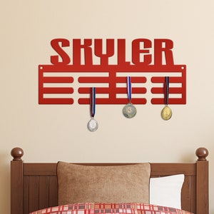 May include: Red metal medal display rack with the name "Skyler" in large letters. The rack has 10 slots for medals and three medals are hanging on the rack. The rack is mounted on a wall above a wooden bed frame with a red and white checked comforter.