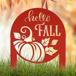 May include: A red metal yard sign with the words "hello FALL" in a decorative font, along with a pumpkin and leaves design. The sign is placed in green grass, with fall foliage in the background.