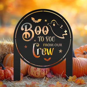 May include: Black metal Halloween yard sign with the words "Boo To You From Our Crew" in a circular design. The sign features a ghost, bats, and stars. Pumpkins and hay bales are in the background.