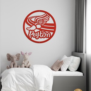 Puede incluir: Un adorno de pared de metal rojo con un dise&ntilde;o estilizado de un pie alado y el nombre "Peyton" en cursiva, dentro de un c&iacute;rculo. La obra de arte est&aacute; montada en una pared blanca sobre una cama con peluches.