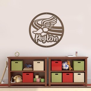Puede incluir: Una pieza de arte mural de metal marr&oacute;n que presenta una zapatilla para correr con alas y el nombre "Peyton" en una fuente cursiva. La obra de arte es circular y cuelga sobre una estanter&iacute;a de madera con cajas de almacenamiento y juguetes coloridos.