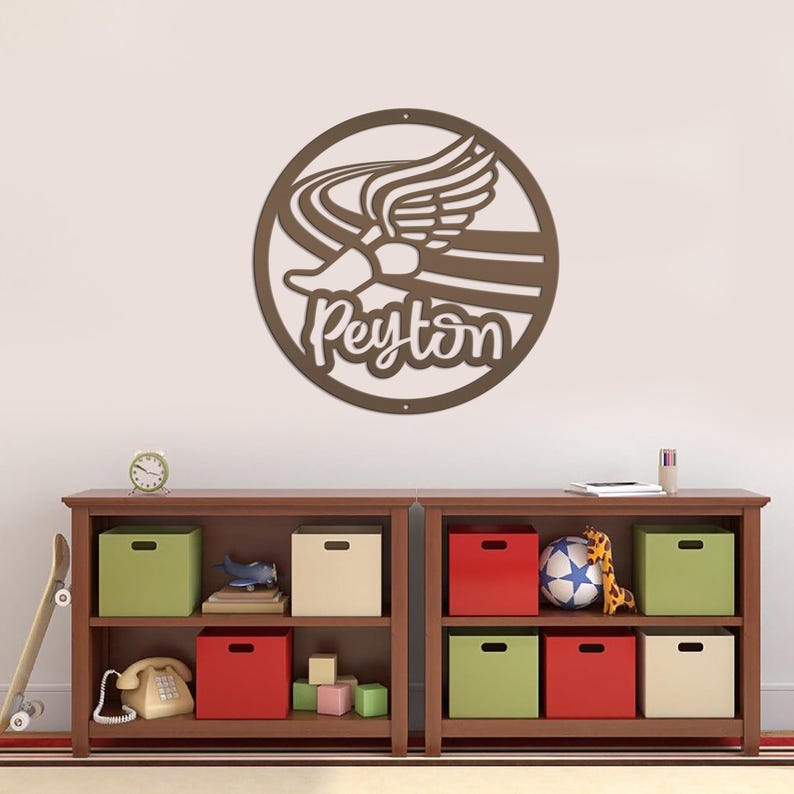 Puede incluir: Una pieza de arte mural de metal marr&oacute;n que presenta una zapatilla para correr con alas y el nombre "Peyton" en una fuente cursiva. La obra de arte es circular y cuelga sobre una estanter&iacute;a de madera con cajas de almacenamiento y juguetes coloridos.