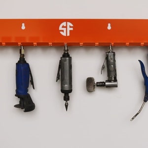 Puede incluir: Un soporte de herramientas de pared naranja con las letras "SF" en un círculo blanco. Cinco herramientas cuelgan del soporte, incluyendo una herramienta neumática azul, una herramienta neumática gris, una herramienta neumática gris con cabeza redonda y una herramienta neumática azul con mango curvo.