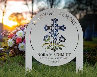 May include: A circular metal memorial garden stake featuring the text IN LOVING MEMORY, a decorative cross, and floral motifs. The name NORA M SCHMIDT and the dates FEB 28, 1934 - MAR 23, 2008 are engraved below the cross with a small scroll design.