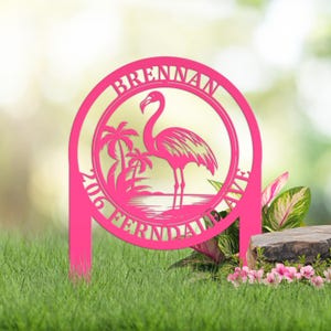 May include: A circular hot pink metal yard sign featuring a cutout silhouette of a flamingo standing near palm trees. The text BRENNAN 206 FERNDALE AVE is integrated into the circular border. The sign has two vertical stakes at the base.