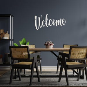 May include: A dining room scene featuring a wooden table and four black chairs with woven backs. A white "Welcome" sign is mounted on the dark blue wall. A small floral arrangement sits on the table, and a rug is on the floor.
