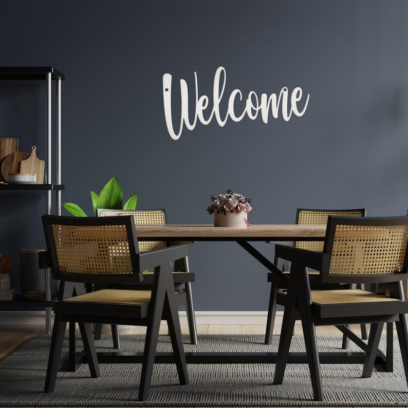 May include: A dining room scene featuring a wooden table and four black chairs with woven backs. A white "Welcome" sign is mounted on the dark blue wall. A small floral arrangement sits on the table, and a rug is on the floor.