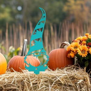 May include: A teal metal garden gnome decoration with a tall hat featuring leaf cutouts. The gnome is placed in front of pumpkins and flowers on a bed of hay, creating a fall harvest scene. The background is blurred with a natural setting.