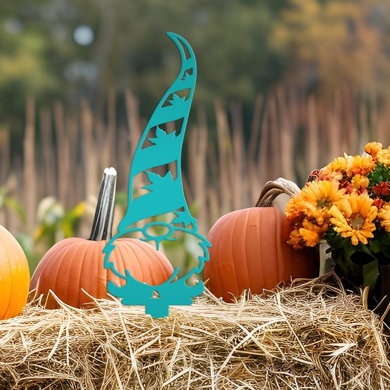 May include: A teal metal garden gnome decoration with a tall hat featuring leaf cutouts. The gnome is placed in front of pumpkins and flowers on a bed of hay, creating a fall harvest scene. The background is blurred with a natural setting.