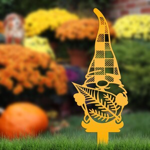 May include: A yellow metal garden gnome stake with a plaid hat and leaf details. The gnome is standing in green grass with a blurred background of orange and yellow flowers.
