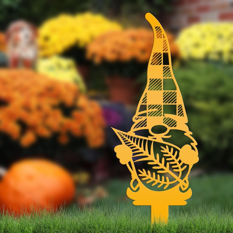 May include: A yellow metal garden gnome stake with a plaid hat and leaf details. The gnome is standing in green grass with a blurred background of orange and yellow flowers.
