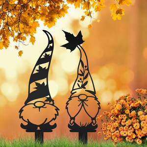 May include: Two black metal garden gnomes with tall, pointed hats. One hat has diagonal stripes and leaf cutouts, the other has a leaf at the top. They are set in a grassy area with an orange and yellow background.