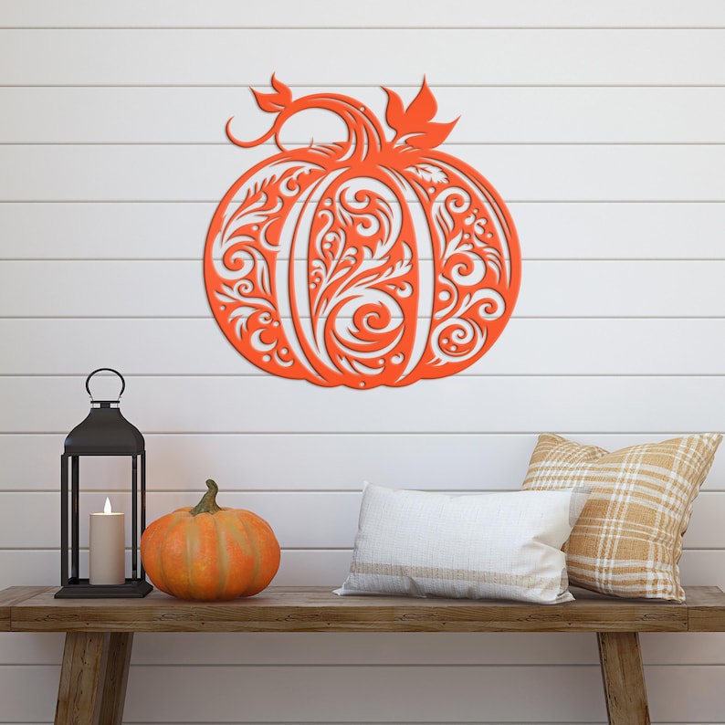 May include: An orange, laser-cut pumpkin decoration with intricate floral patterns, displayed on a white shiplap wall. The pumpkin is the focal point, complemented by a small pumpkin, a candle lantern, and decorative pillows on a wooden bench, creating a fall-themed display.