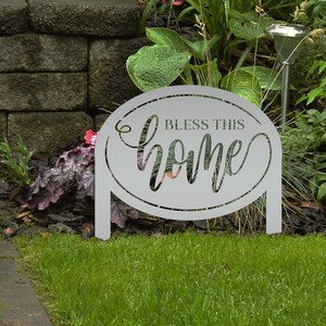 Puede incluir: Un letrero de jardín de metal gris con las palabras "Bless This Home" recortadas en una fuente decorativa. El letrero tiene forma ovalada y tiene dos patas para colocarlo en un jardín. El letrero está rodeado de hierba verde y plantas.
