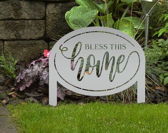 Puede incluir: Un letrero de jardín de metal gris con las palabras "Bless This Home" recortadas en una fuente decorativa. El letrero tiene forma ovalada y tiene dos patas para colocarlo en un jardín. El letrero está rodeado de hierba verde y plantas.