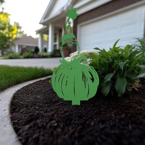 May include: A green metal garden stake featuring a gnome with a pumpkin body. The gnome wears a striped hat with a leaf accent. The stake is placed in a garden bed with dark mulch and green plants.