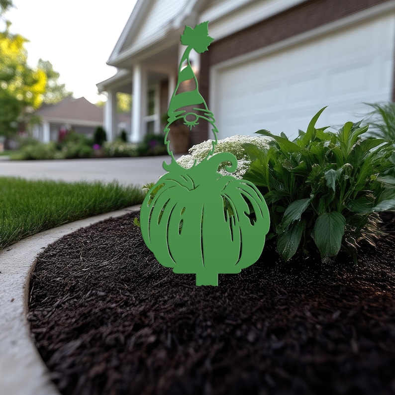 May include: A green metal garden stake featuring a gnome with a pumpkin body. The gnome wears a striped hat with a leaf accent. The stake is placed in a garden bed with dark mulch and green plants.