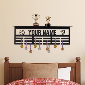 May include: Black metal sports medal display with the text "YOUR NAME", a baseball and a football. Includes a trophy, a star-shaped award, and several medals hanging below. The medals have purple and blue ribbons.