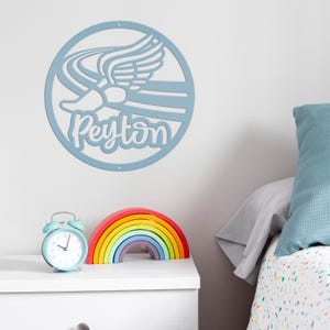 Puede incluir: Adorno de pared azul claro con un dise&ntilde;o de zapato alado y el nombre "Peyton" en cursiva. Un arco&iacute;ris de madera y un despertador azul claro est&aacute;n sobre una mesita de noche blanca. Una almohada azul descansa sobre una cama.